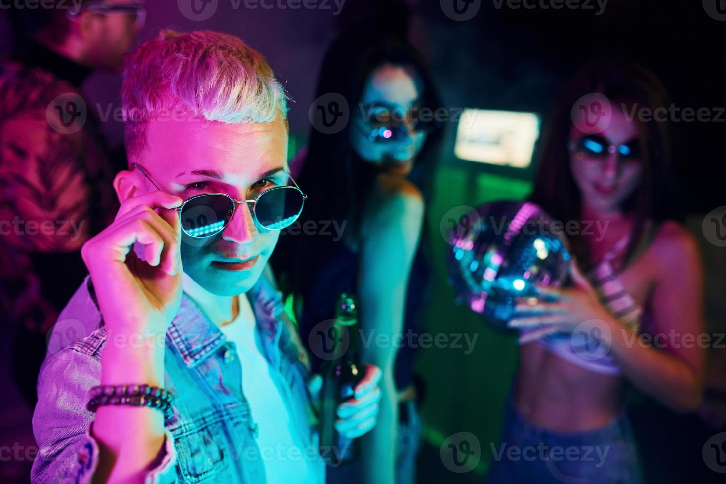 Hipster guy in sunglasses and with bottle of alcohol posing for camera in front of young people that having fun in night club with colorful laser lights photo