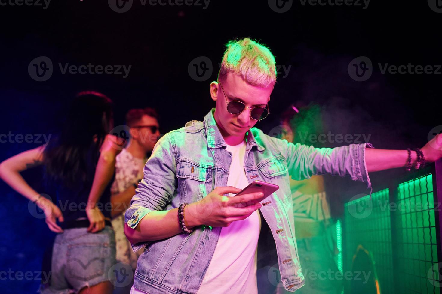 chico parado con un teléfono inteligente frente a jóvenes que se divierten en un club nocturno con luces láser de colores foto