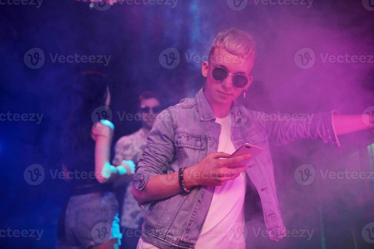 Guy standing with smartphone in front of oung people that having fun in night club with colorful laser lights photo