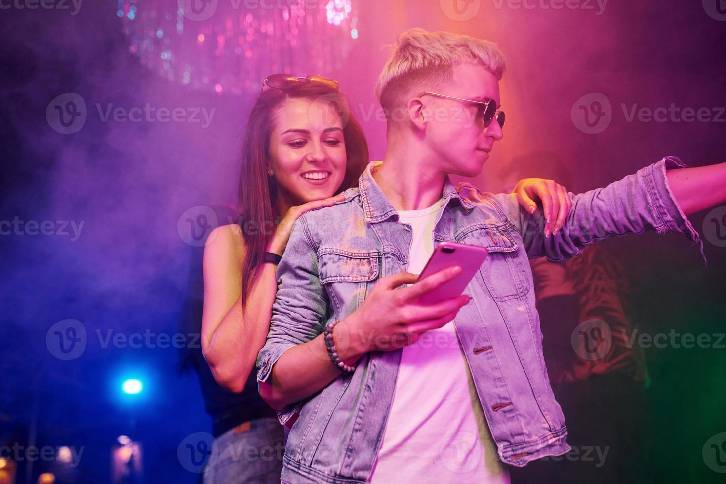 chico parado con un teléfono inteligente frente a jóvenes que se divierten en un club nocturno con luces láser de colores foto