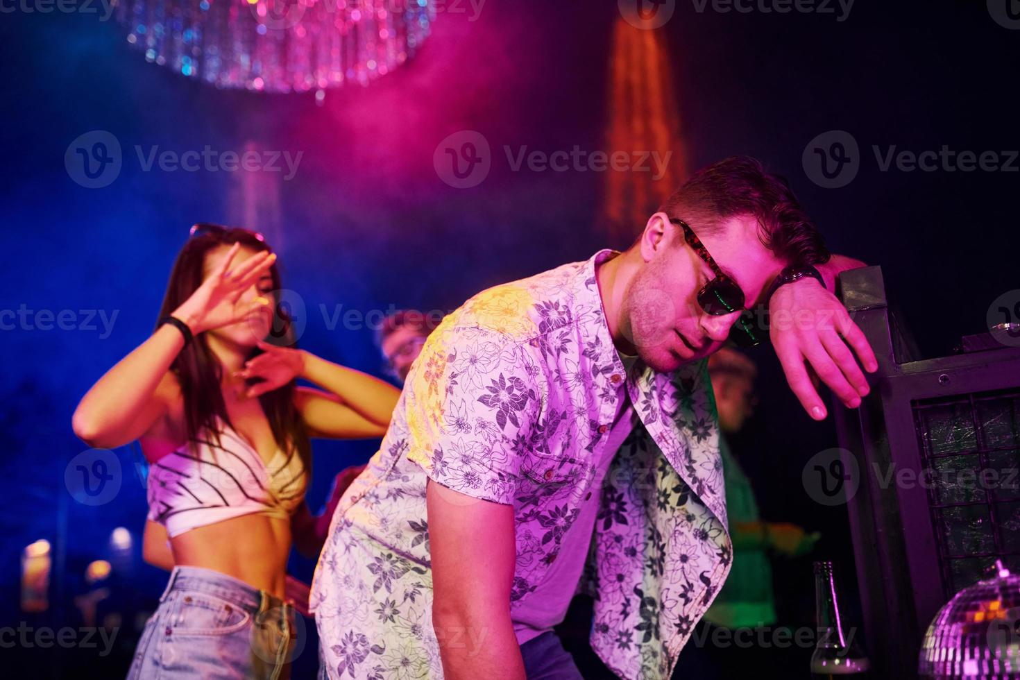 borracho apoyado en la cerca frente a los jóvenes que se divierten en el club nocturno con luces láser de colores foto