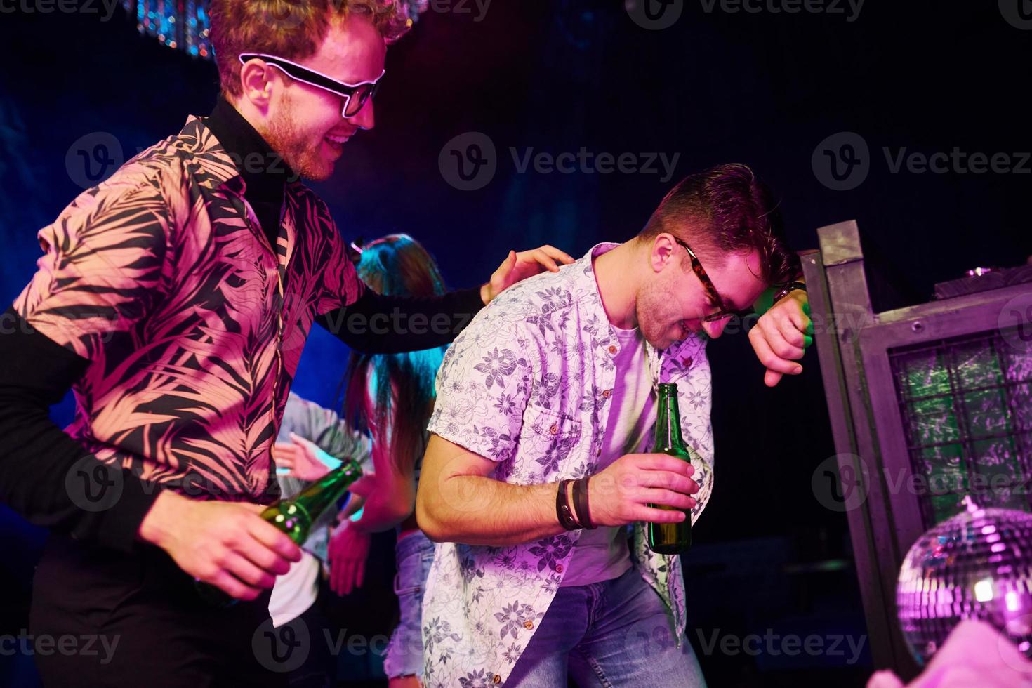 Drunk guy leaning in fence in front of young people that having fun in night club with colorful laser lights photo