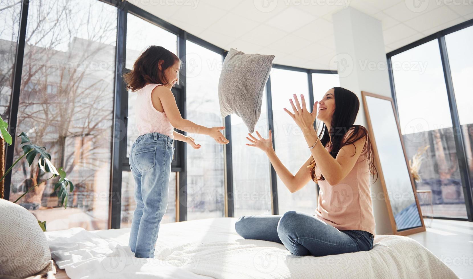 divirtiéndose con la almohada en la cama. joven madre con su hija pasando el fin de semana juntos en la habitación foto