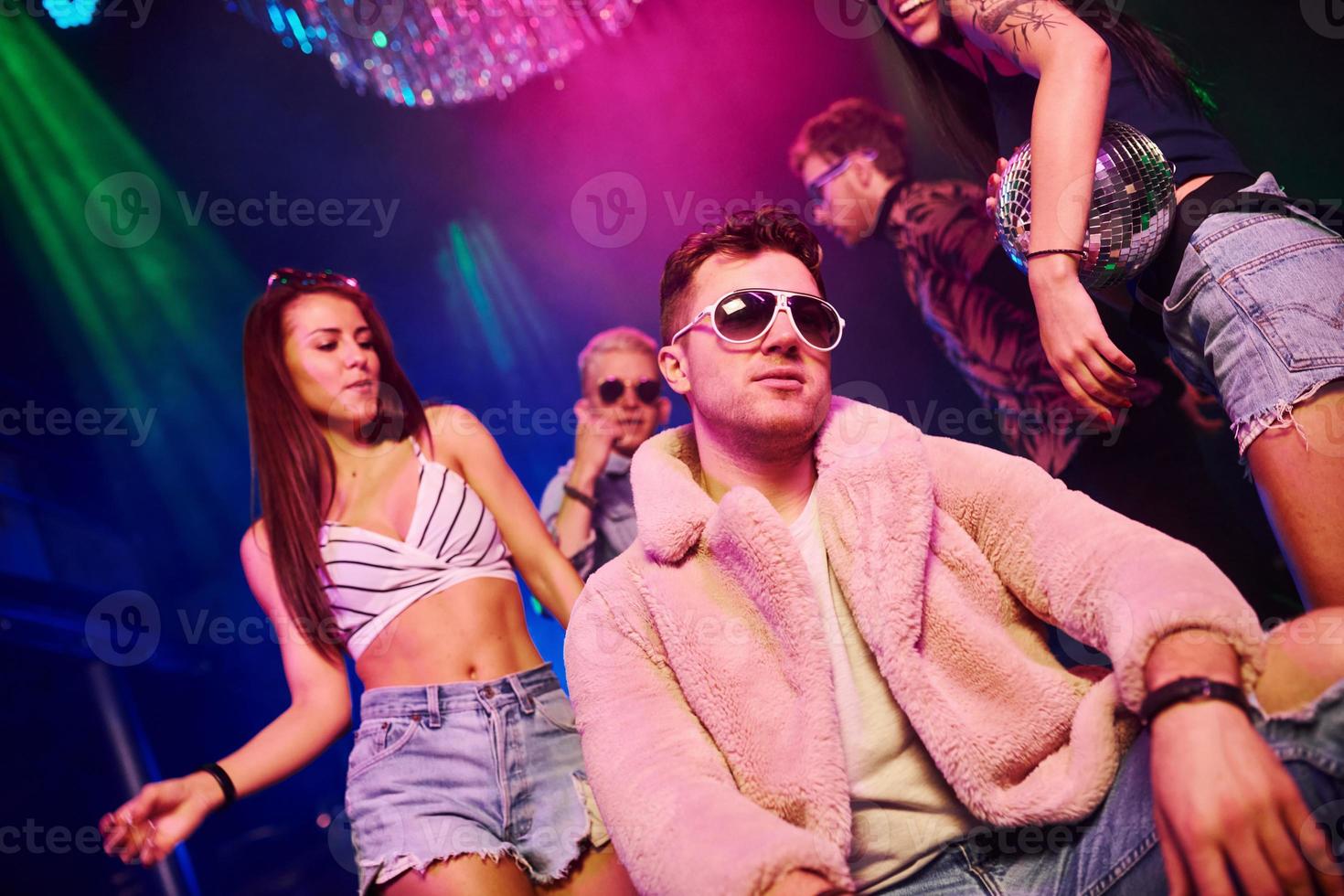 Elegant man in sunglasses in the midle feels like a boss. Young people is having fun in night club with colorful laser lights photo