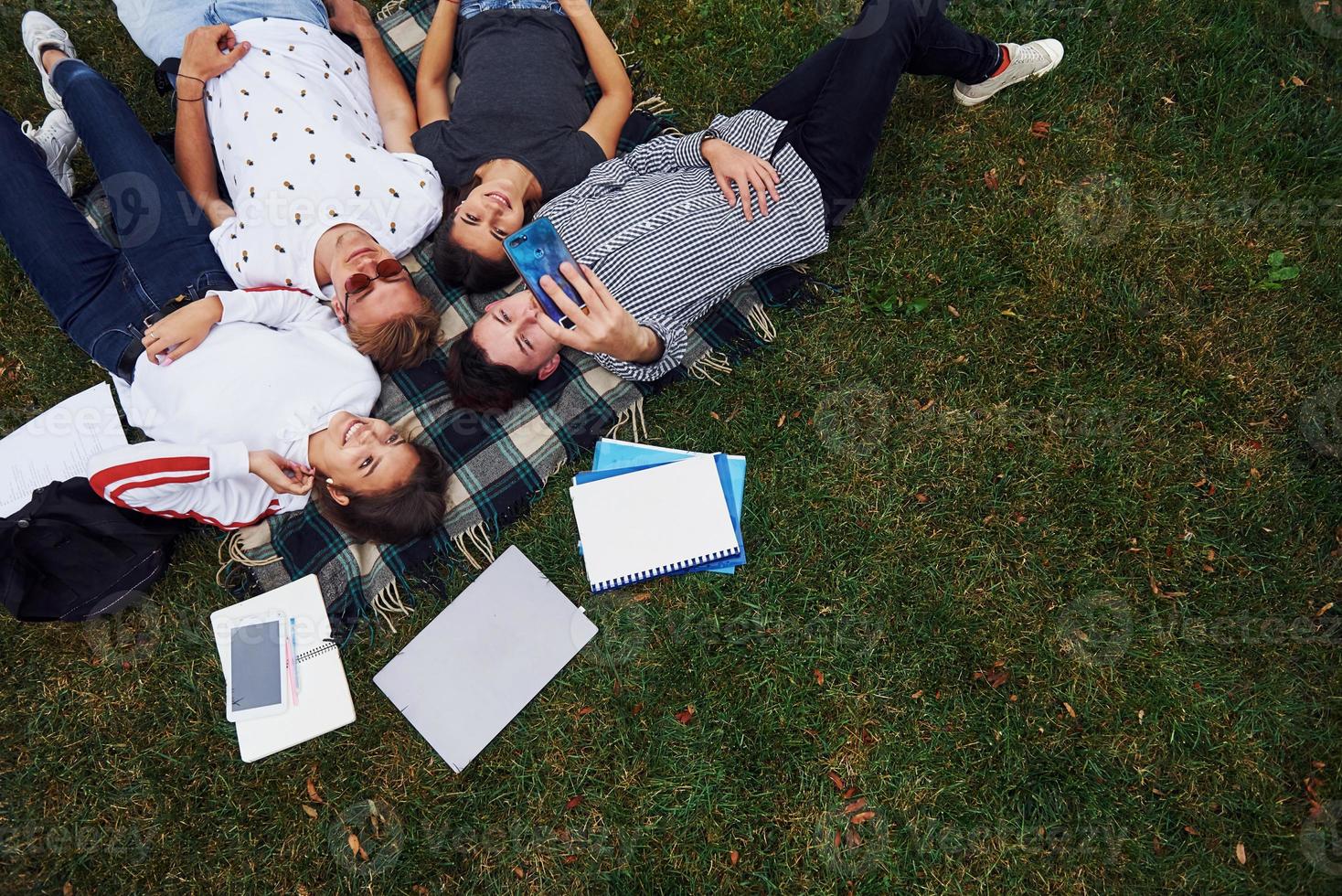 Taking selfie by using smartphone. Group of young students in casual clothes on green grass at daytime photo