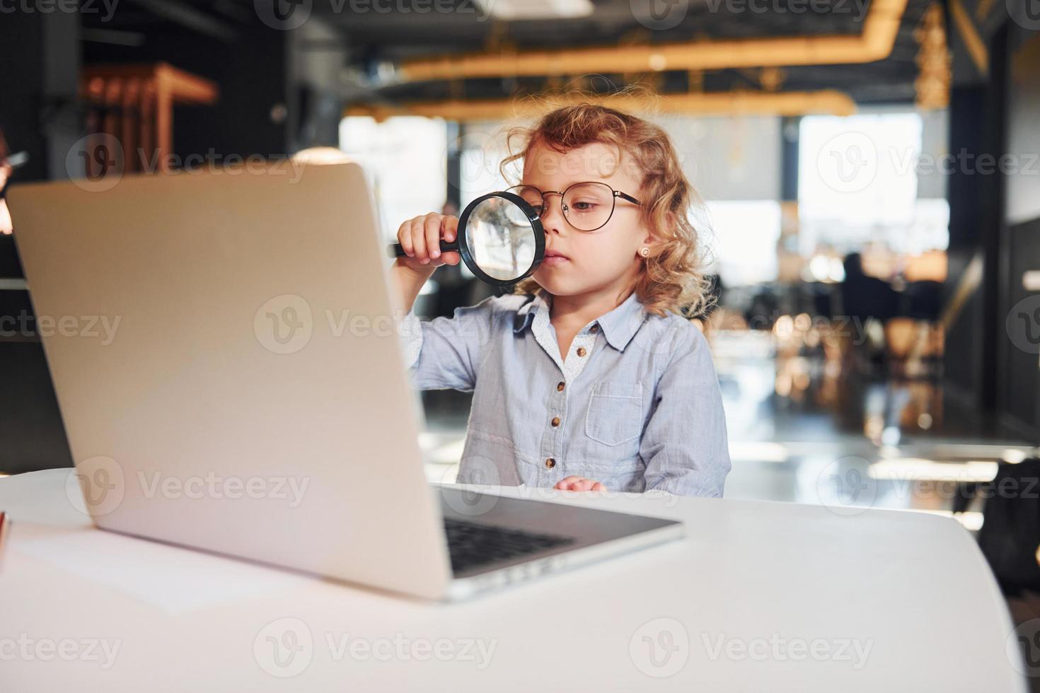 Smart child in casual clothes with laptop on table have fun with magnifying glass photo