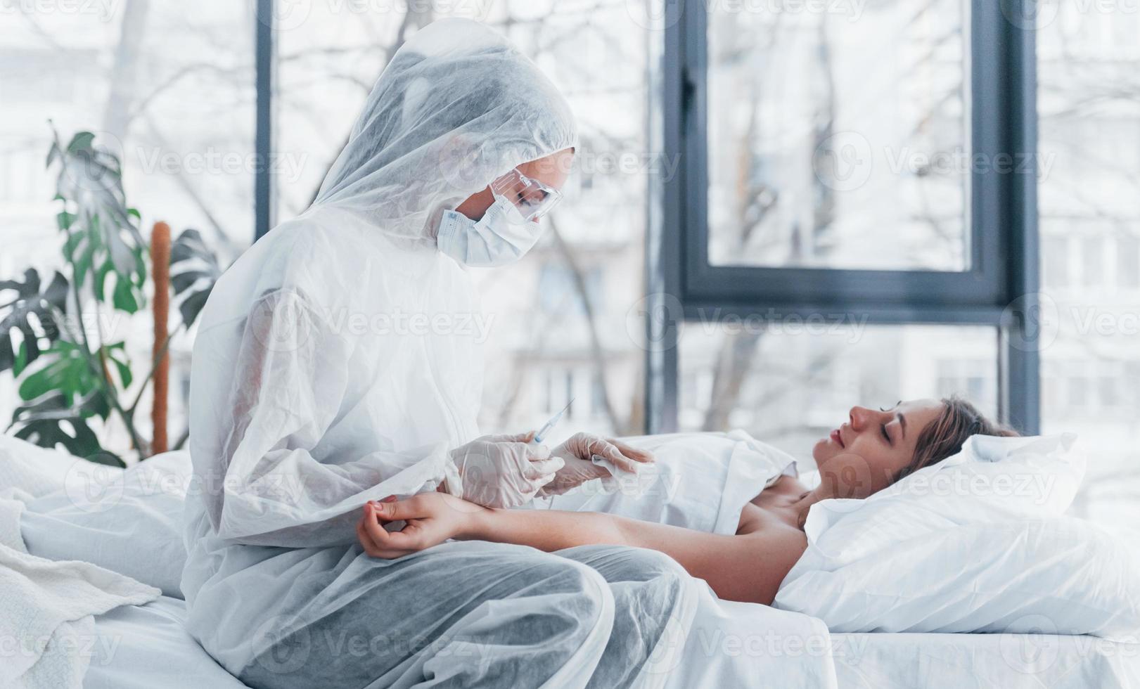 Female doctor in defensive lab coat and protective eyewear with syringe in hand injecting medicine to young girl sick of virus photo