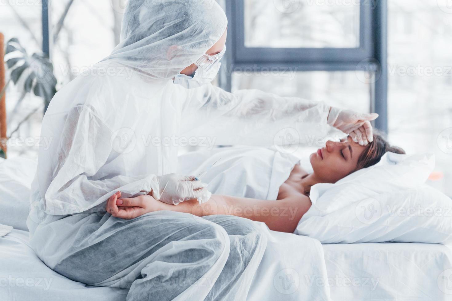 comprueba la temperatura. doctora en bata de laboratorio defensiva y anteojos protectores con jeringa en la mano inyectando medicamentos a una joven enferma de virus foto