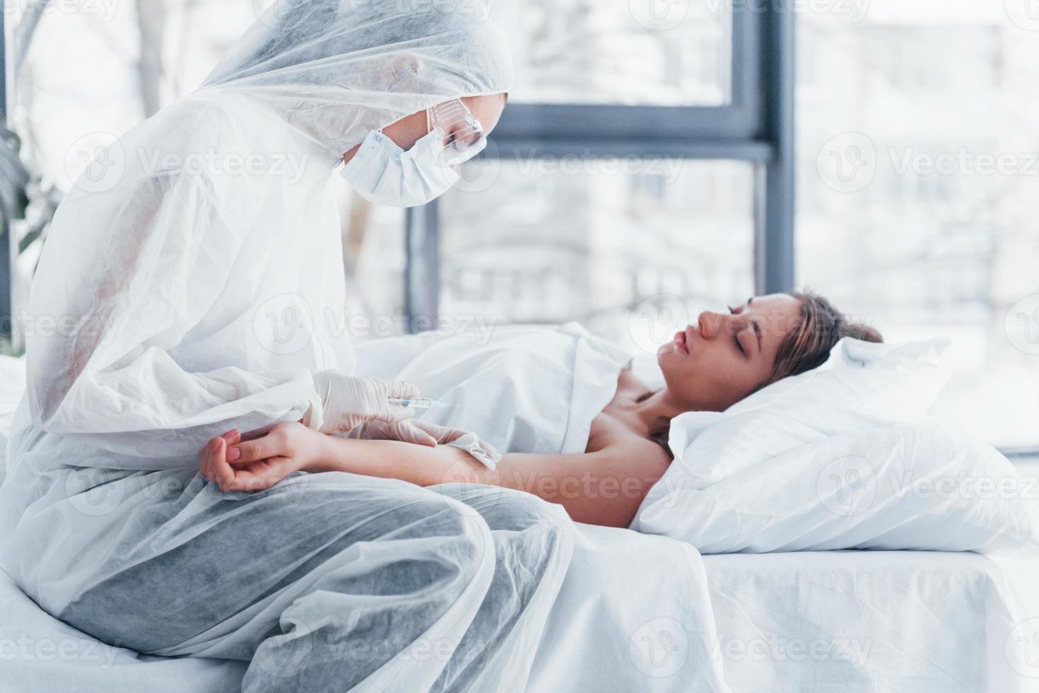 Female doctor in defensive lab coat and protective eyewear with syringe in hand injecting medicine to young girl sick of virus photo