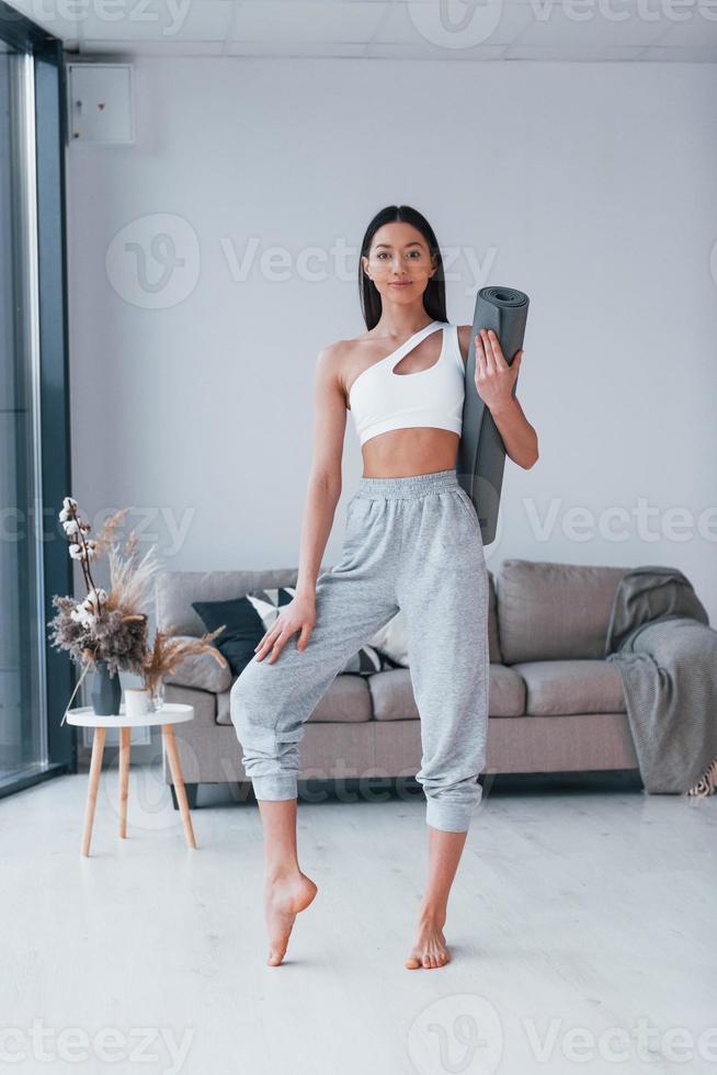 Standing and holds mat. Young woman with slim body shape in sportswear have fitness day indoors at home photo