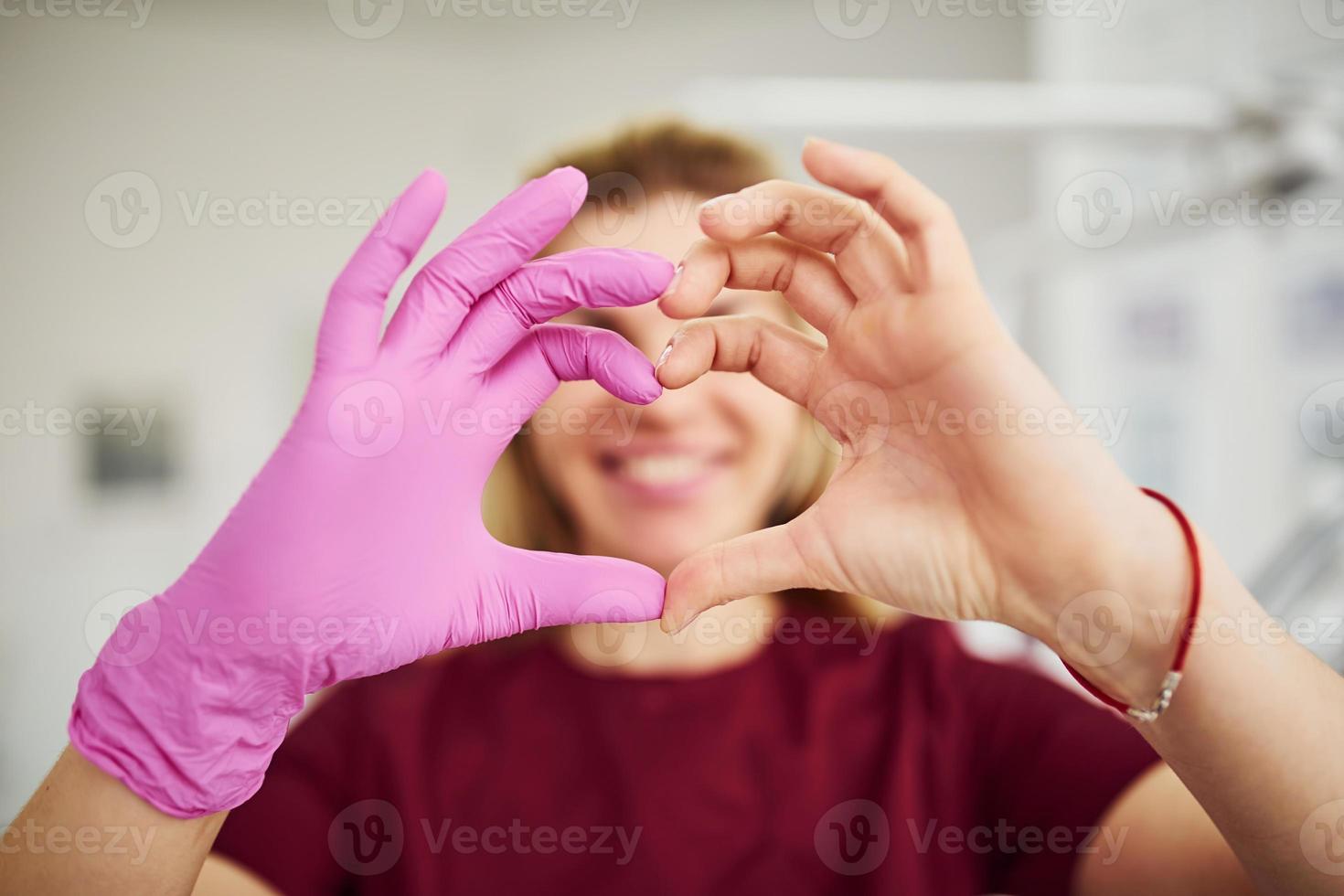 joven dentista uniformada parada en la oficina de estomatología y haciendo gestos en forma de corazón foto