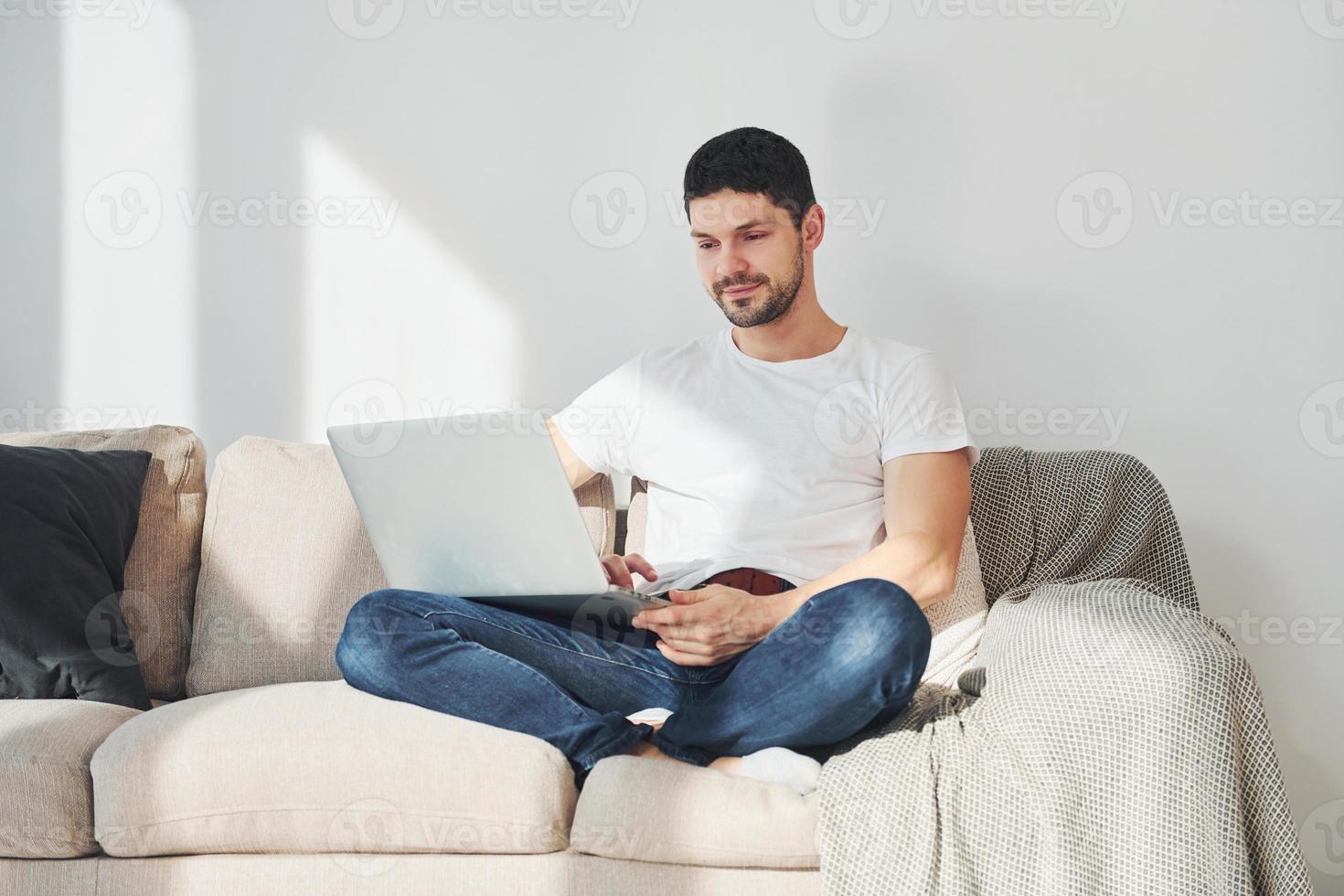 Man in white shirt and jeans sitting on bed with laptop indoors photo