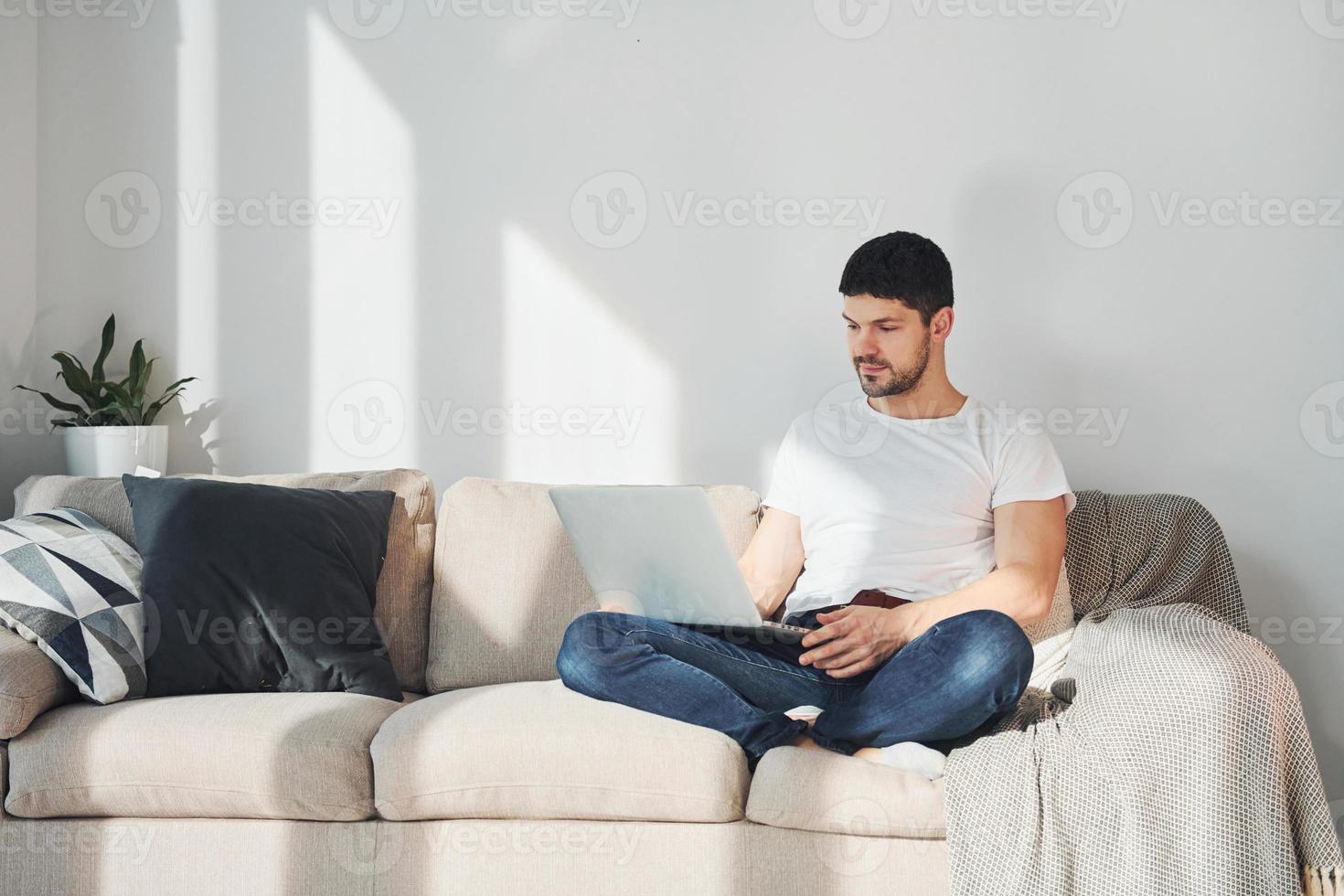 Man in white shirt and jeans sitting on bed with laptop indoors photo