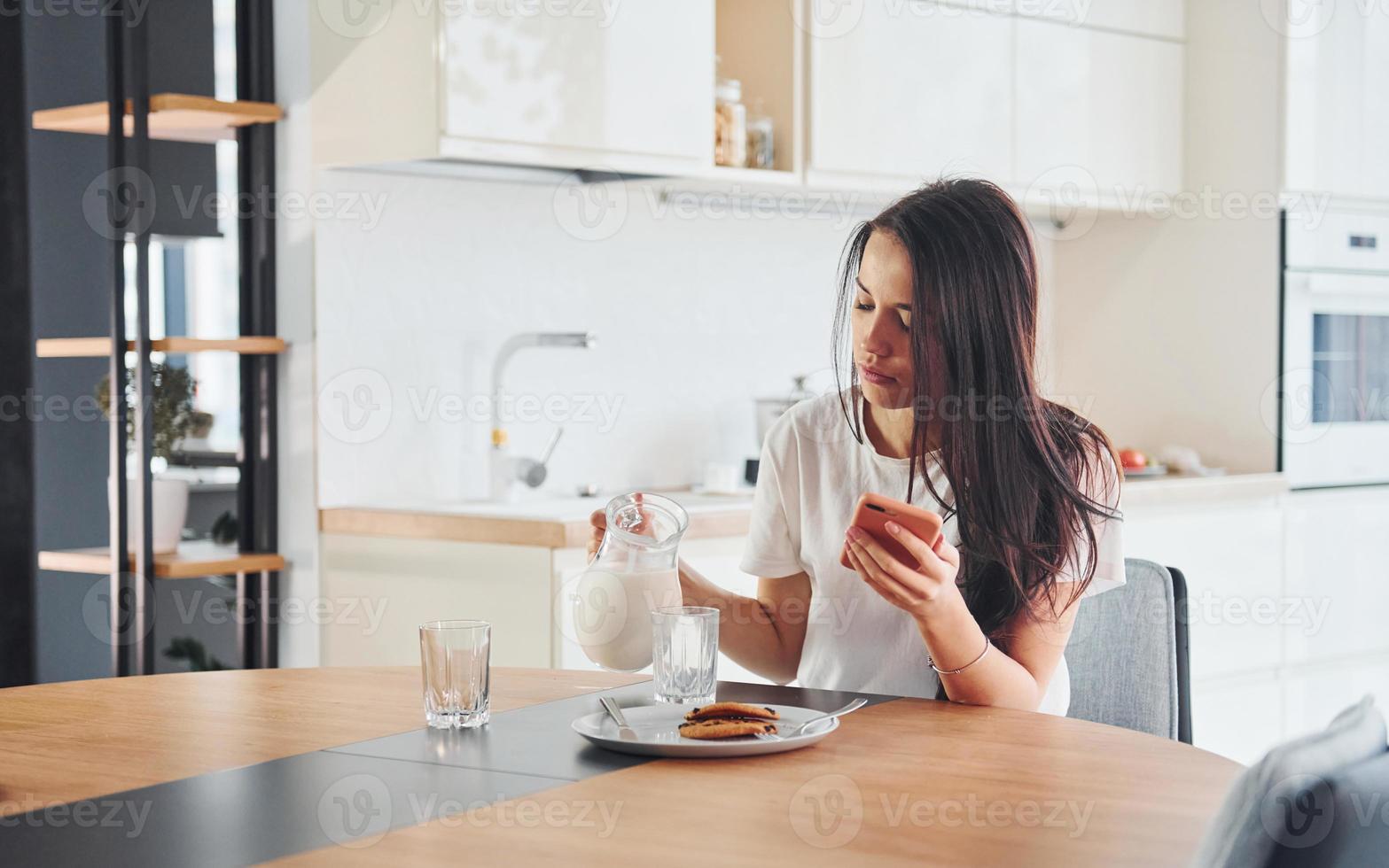 Pouring fresh milk into the glass. Young beautiful brunette in casual clothes indoors in kitchen at daytime photo