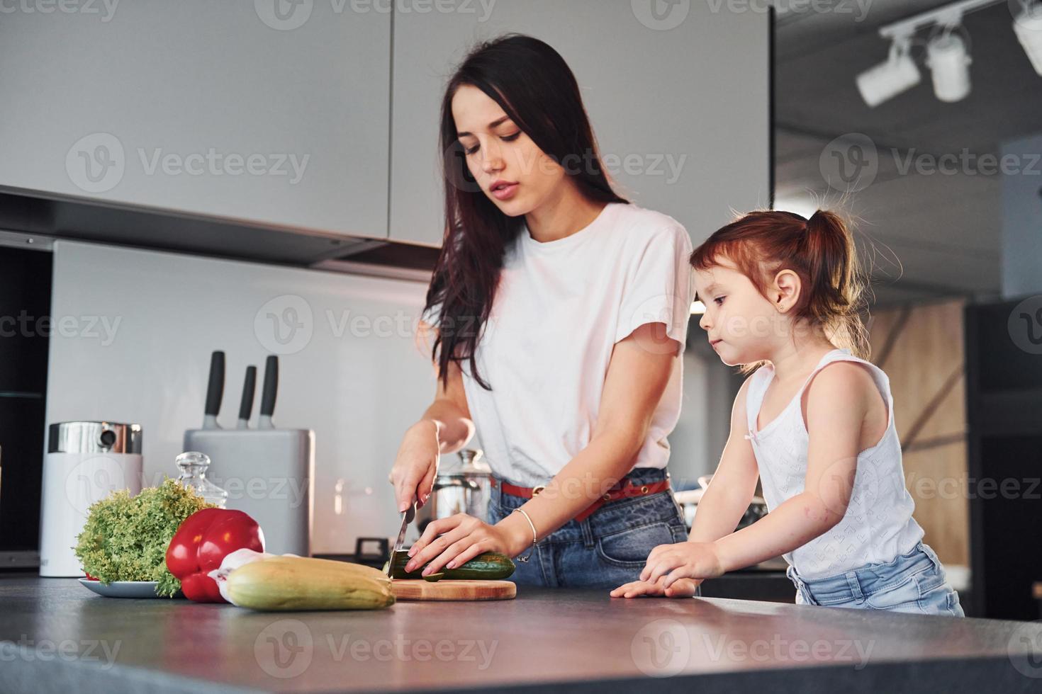 Mother with her little daughter slicing vegetables indoors in kitchen photo