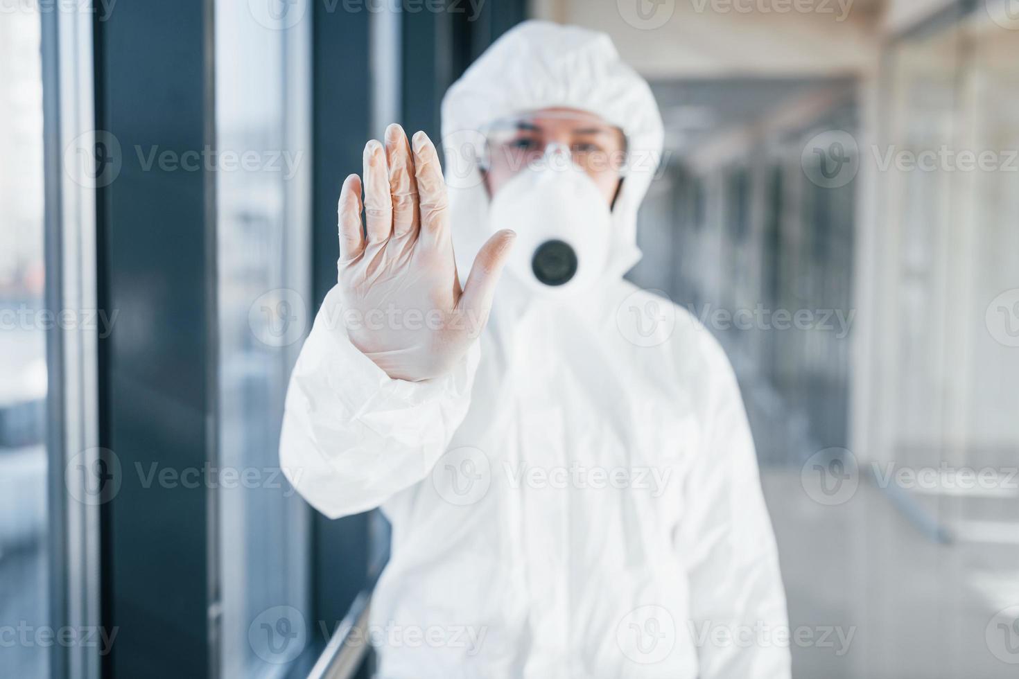 Stop the virus gesture. Female doctor scientist in lab coat, defensive eyewear and mask standing indoors photo