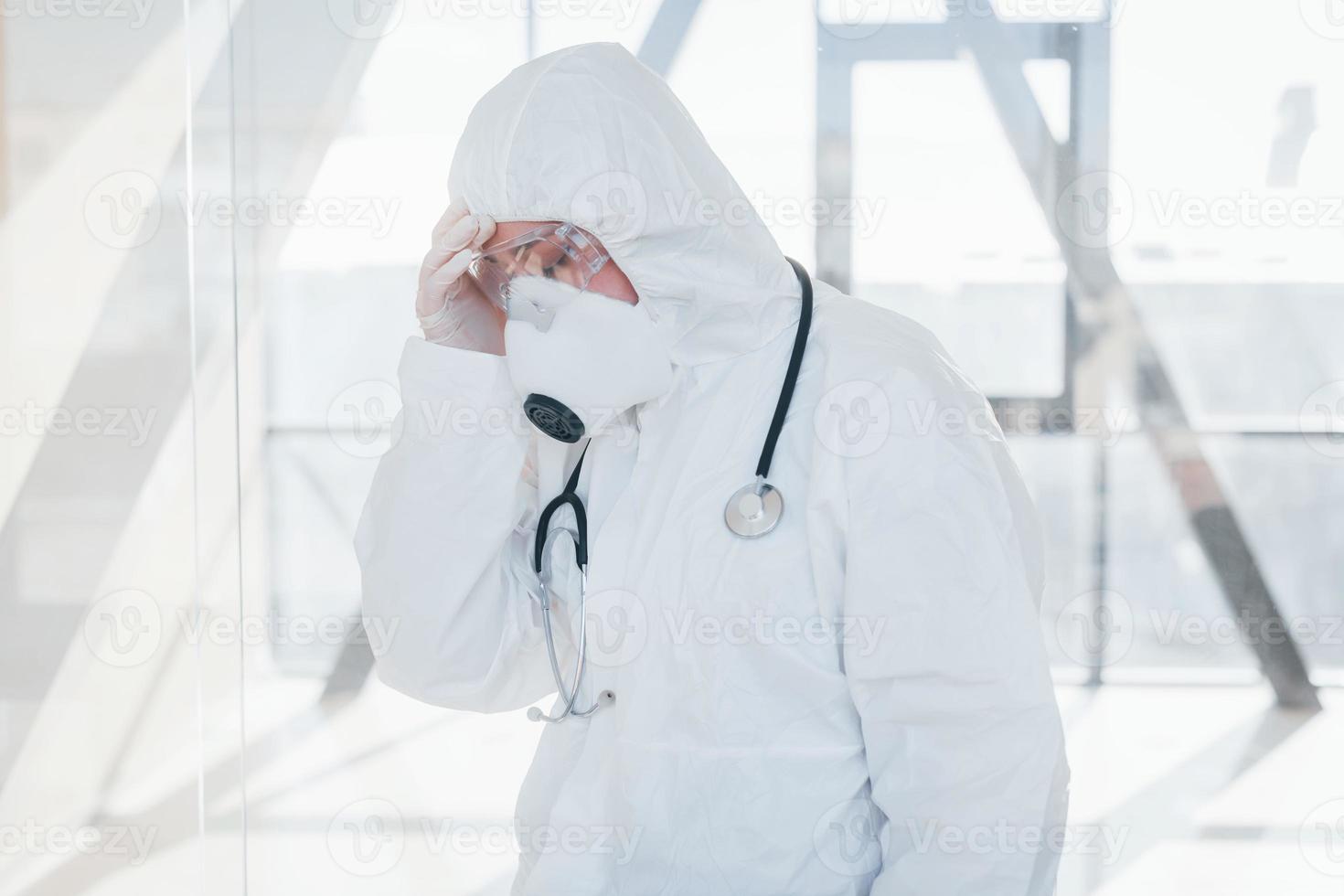 Feels bad and sick. Female doctor scientist in lab coat, defensive eyewear and mask standing indoors photo