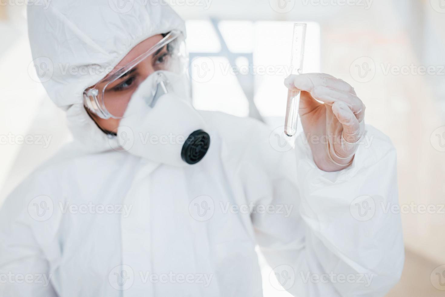 With test tube in hand. Female doctor scientist in lab coat, defensive eyewear and mask standing indoors photo
