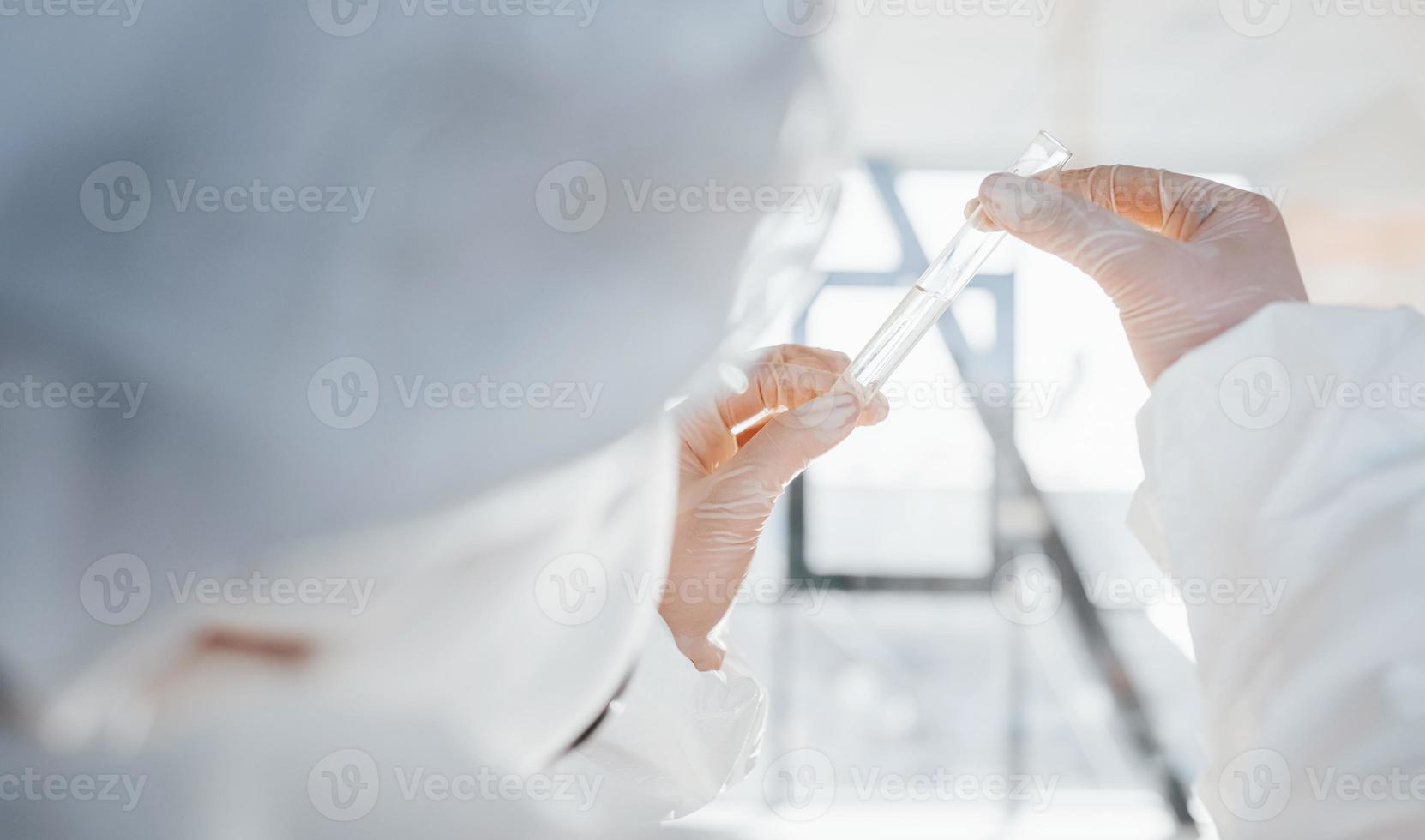 With test tube in hand. Female doctor scientist in lab coat, defensive eyewear and mask standing indoors photo