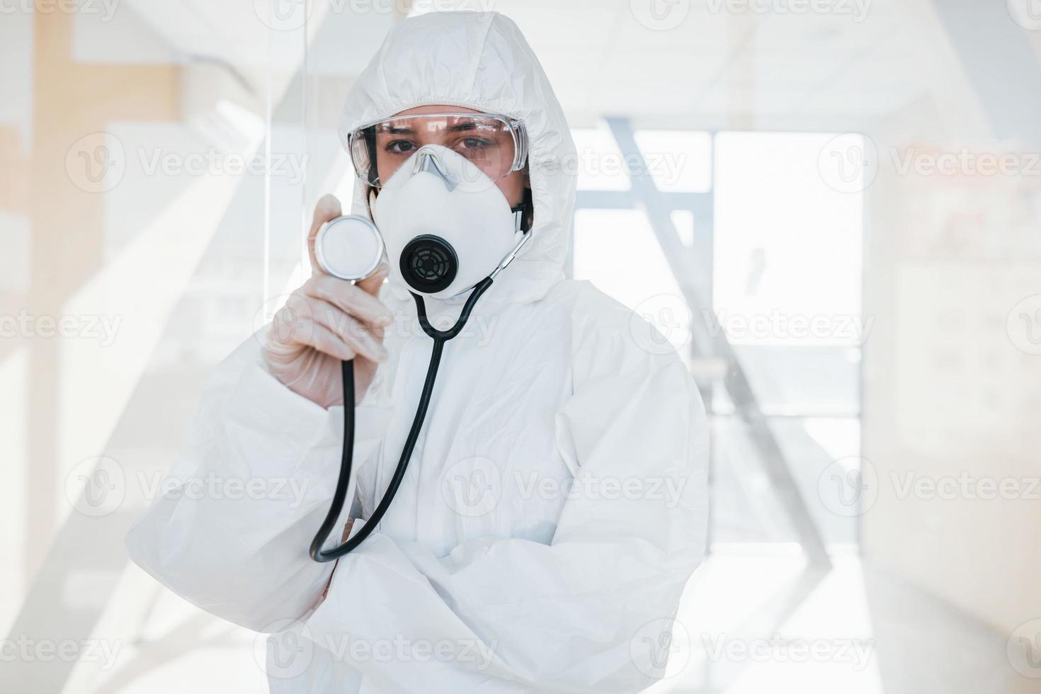 Female doctor scientist in lab coat, defensive eyewear and mask standing indoors and holds stethoscope photo