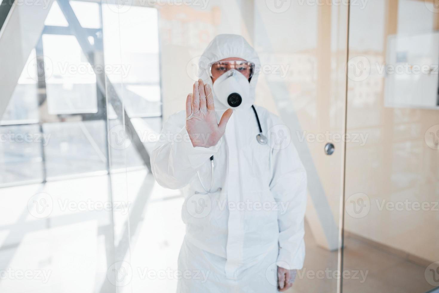 Stop the virus gesture. Female doctor scientist in lab coat, defensive eyewear and mask standing indoors photo