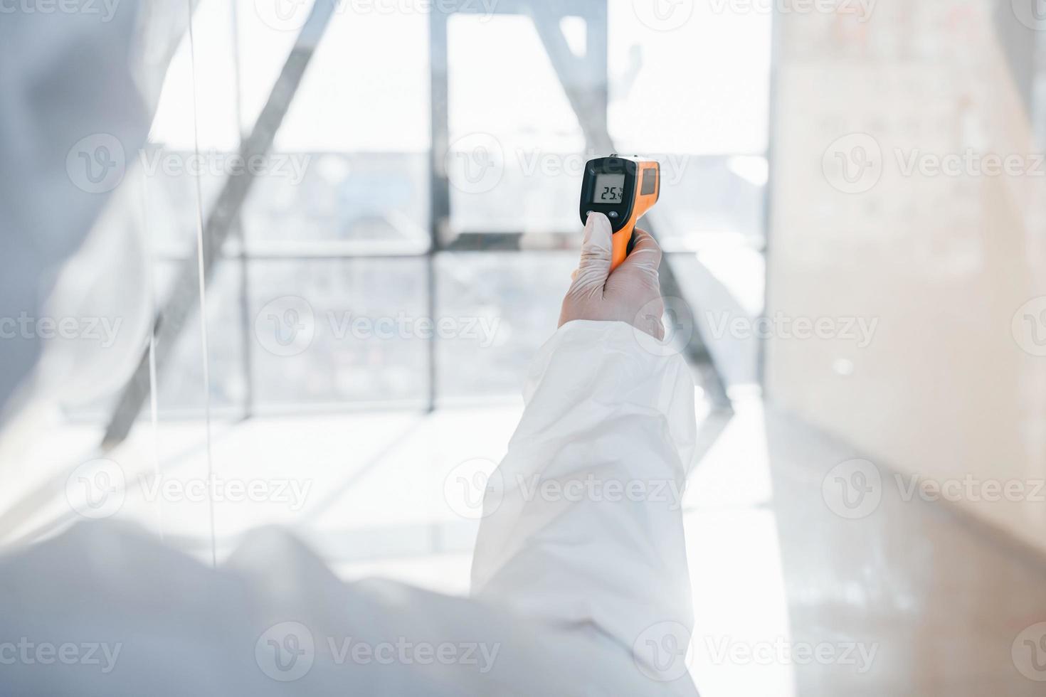 Female doctor scientist in lab coat, defensive eyewear and mask standing indoors with infrated thermometer photo
