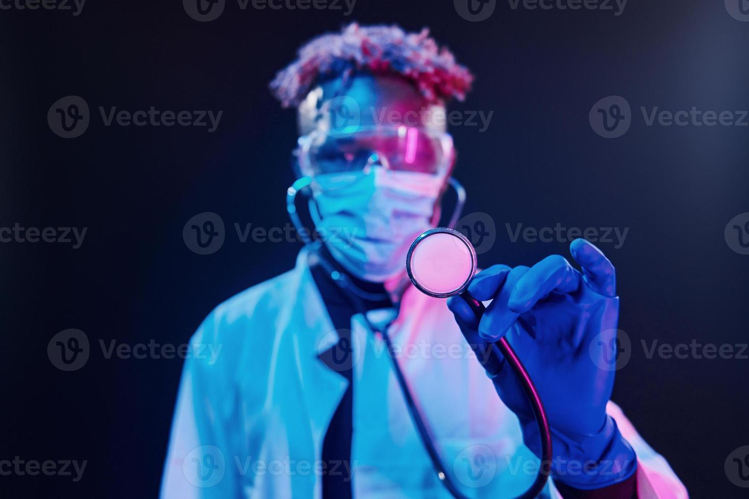 Smart doctor in protective uniform holding stethoscope. Futuristic neon lighting. Young african american man in the studio photo