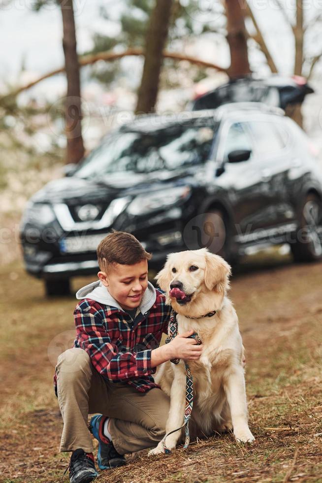 Cheerful boy in casual clothes sitting with her dog in forest against modern black car photo