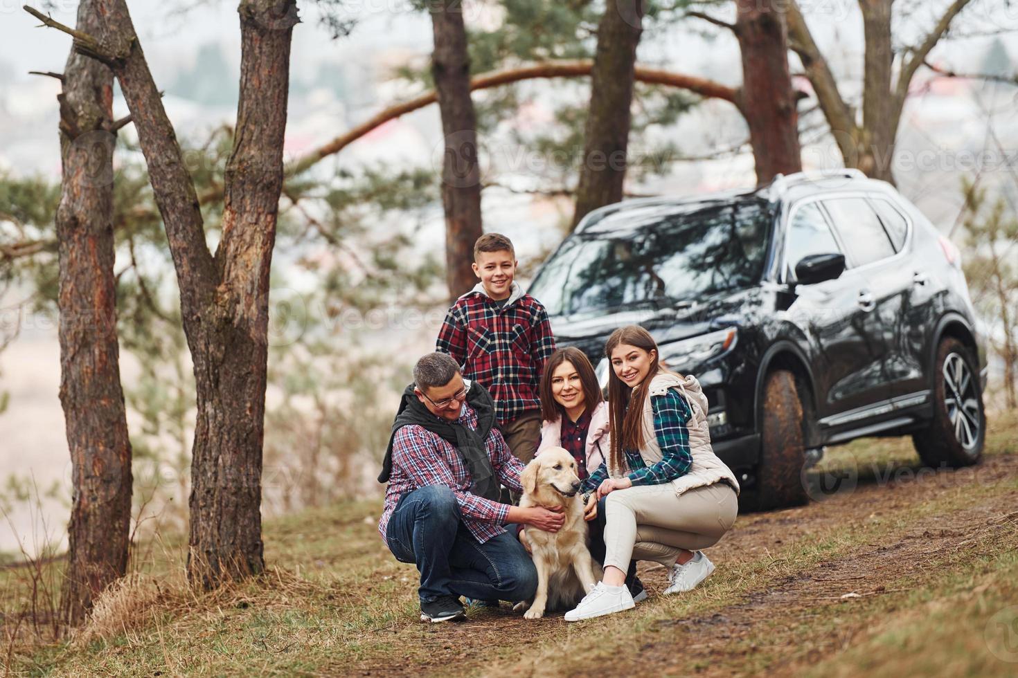 Happy family sitting and having fun with their dog near modern car outdoors in forest photo