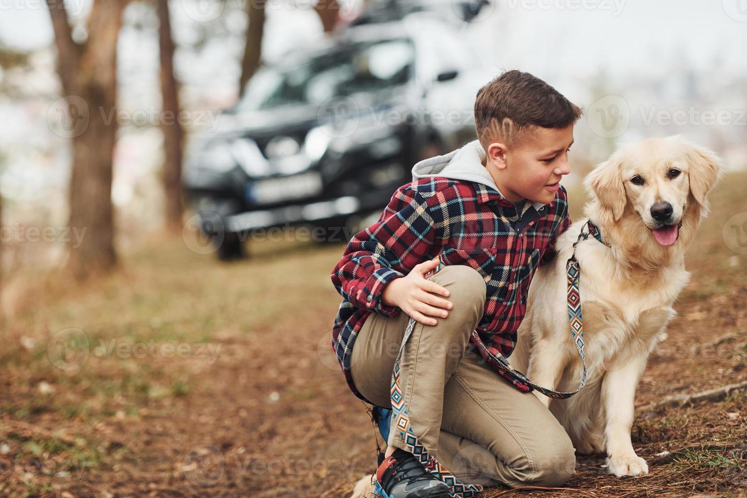 Cheerful boy in casual clothes sitting with her dog in forest against modern black car photo