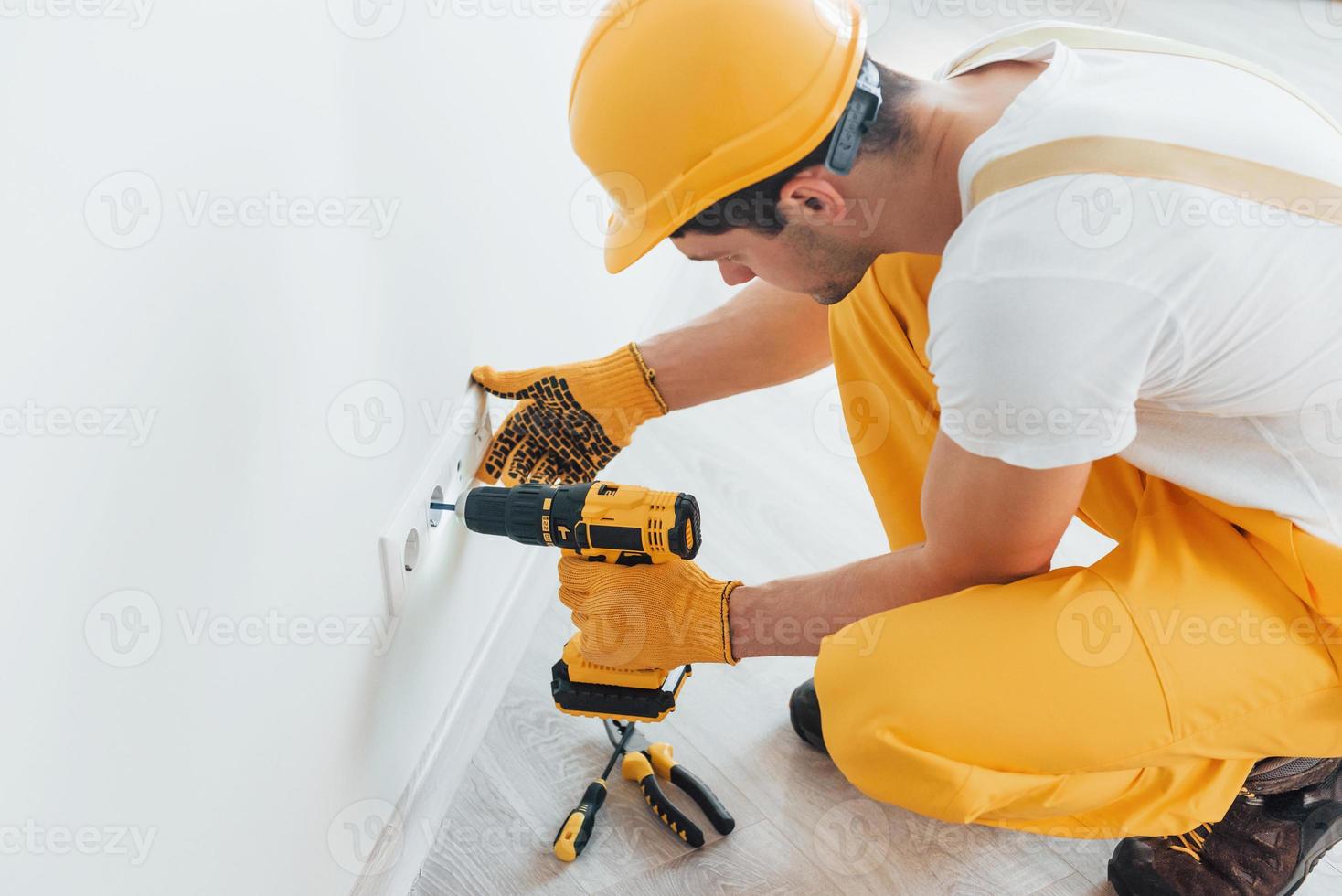 Handyman in yellow uniform works with electricity and installing new socket by using automatic screwdriver. House renovation conception photo