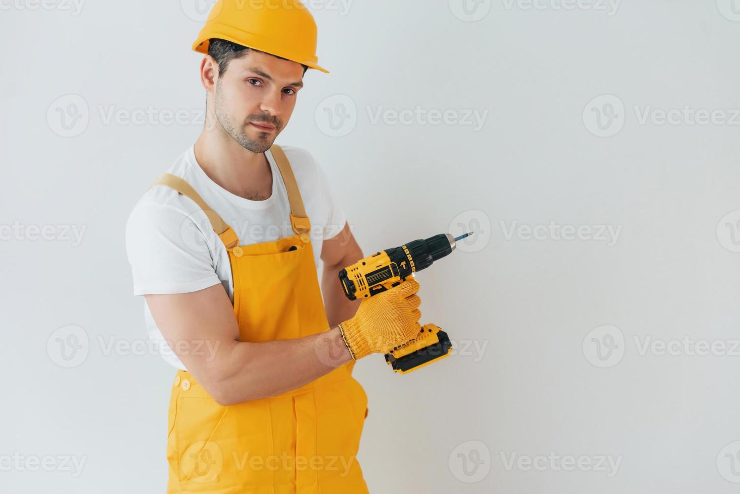 Handyman in yellow uniform standing against white wall with automatic screwdriver. House renovation conception photo