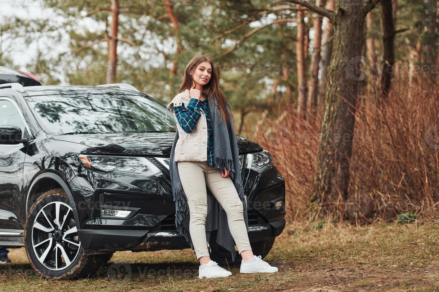 Young cheerful girl standing near modern black car outdoors in the forest photo