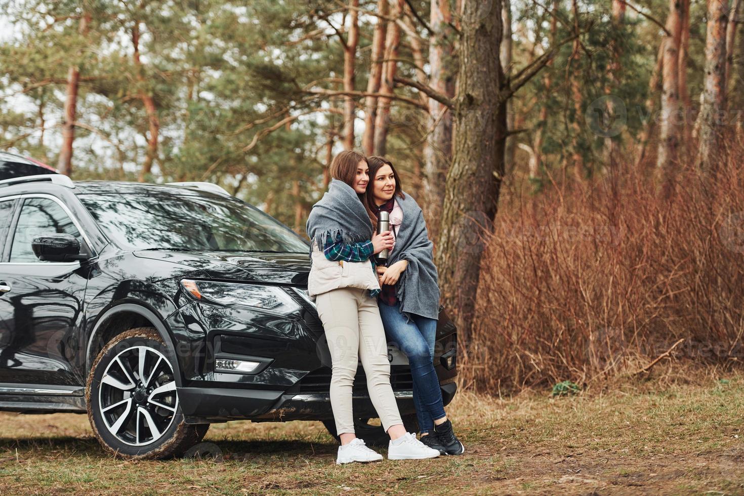 Mother and daughter standing together near modern black car outdoors in the forest photo