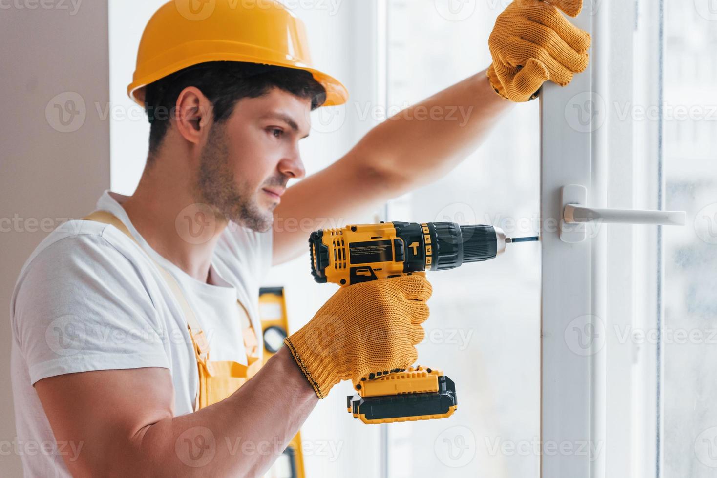Handyman in yellow uniform installs new window by using automatic screwdriver. House renovation conception photo