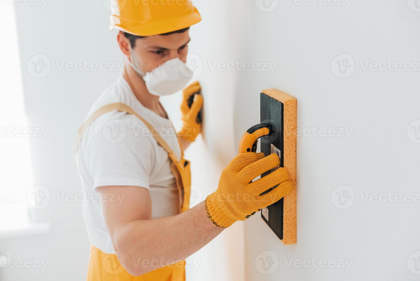 Handyman in yellow uniform and protective mask polishing wall indoors. House renovation conception photo