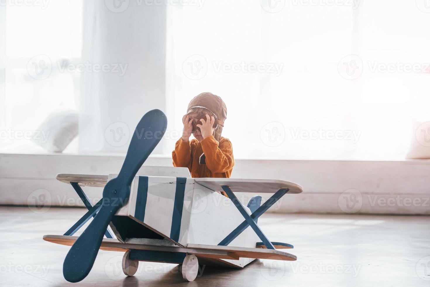 Little boy in retro pilot uniform having fun with toy plane indoors photo