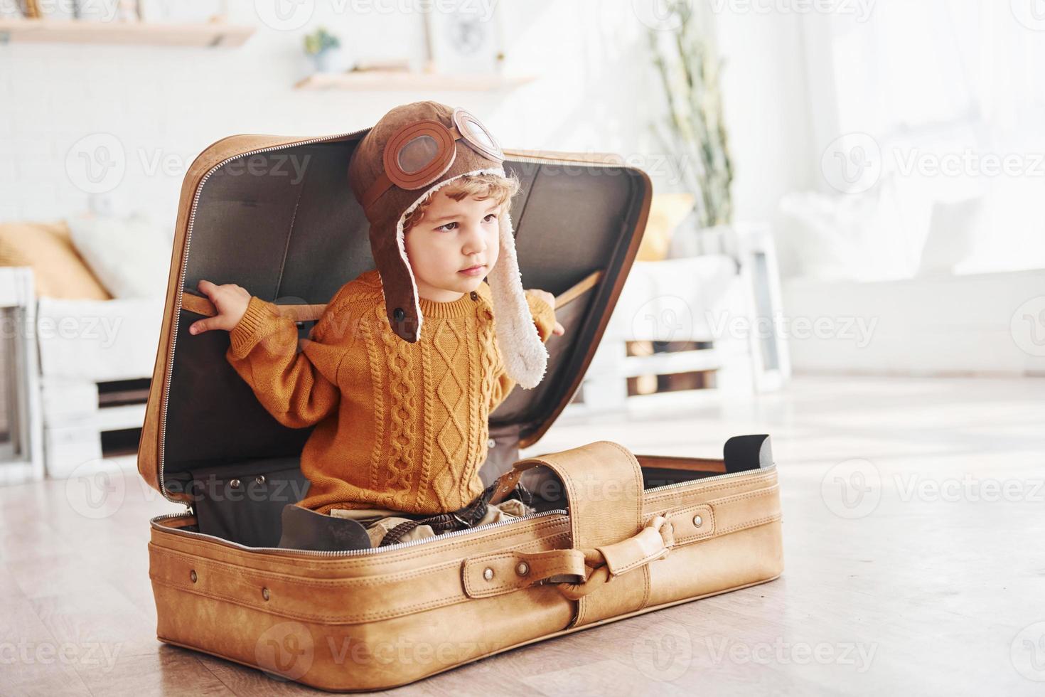 Little boys in retro pilot costume have fun and sitting in suitcase indoors at daytime photo