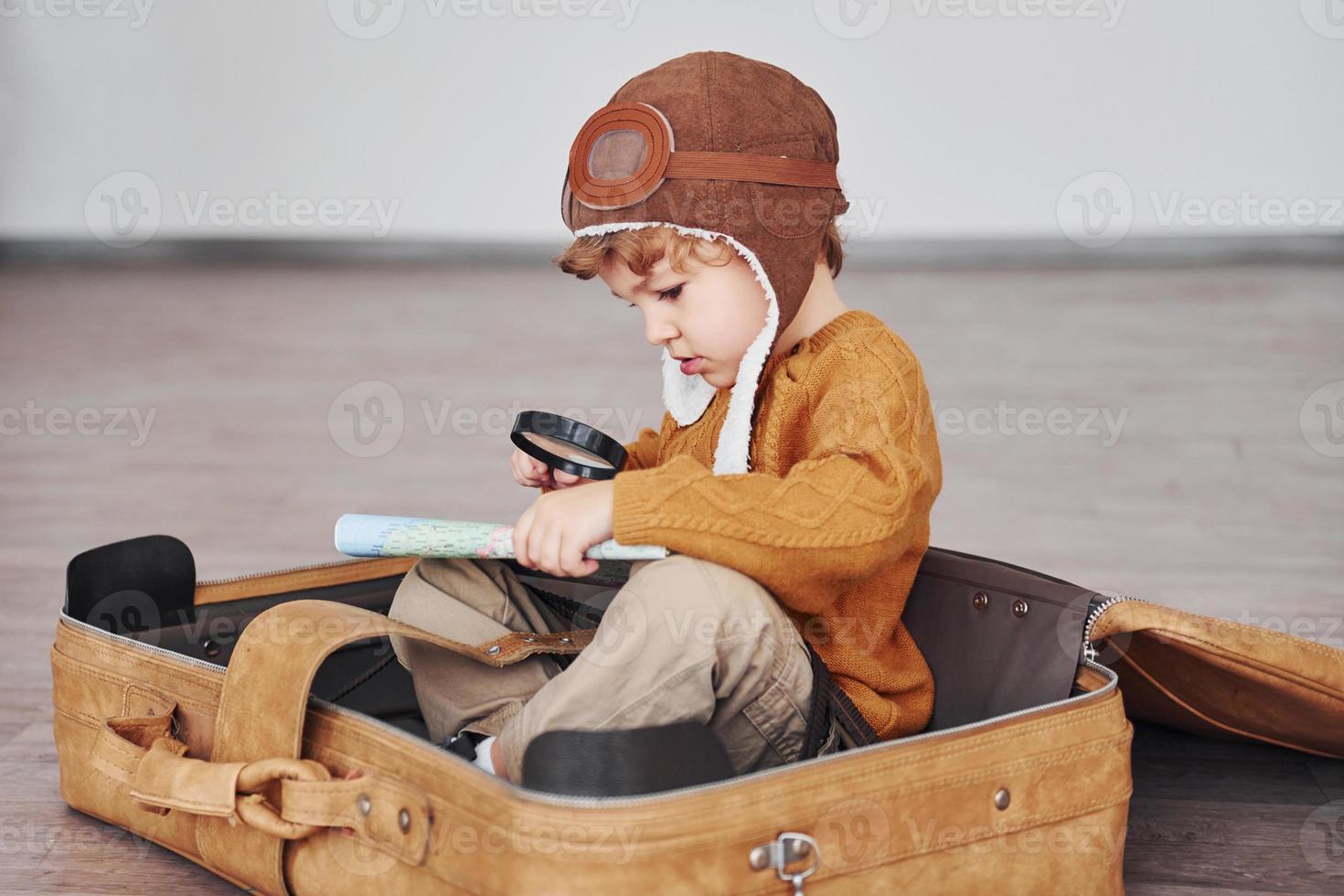 Little boys in retro pilot costume have fun and sitting in suitcase indoors at daytime photo