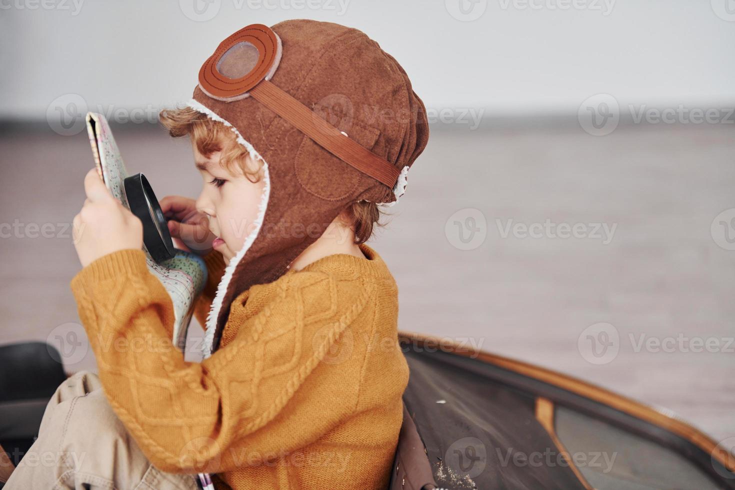 Little boys in retro pilot costume have fun and sitting in suitcase indoors at daytime photo