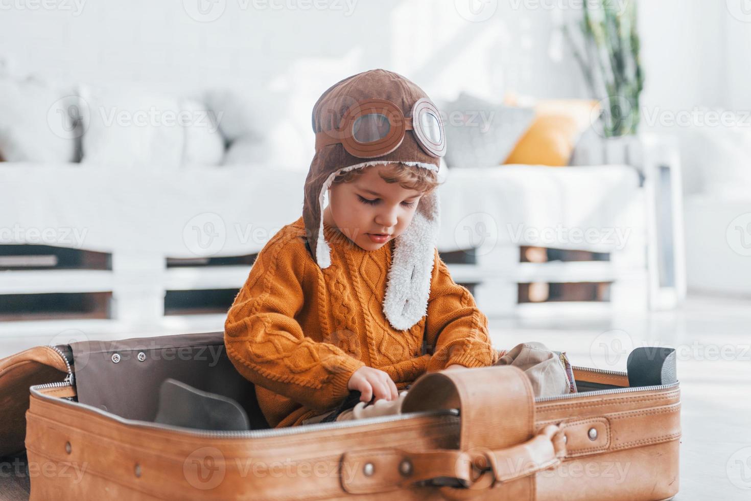 Little boys in retro pilot costume have fun and sitting in suitcase indoors at daytime photo