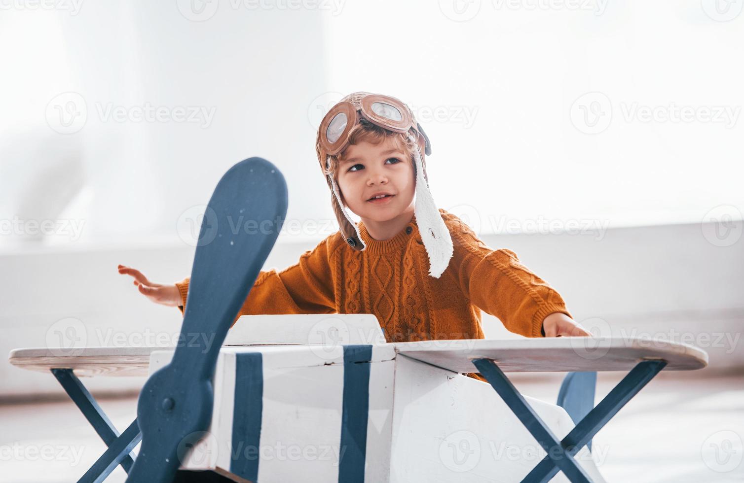 Little boy in retro pilot uniform having fun with toy plane indoors photo