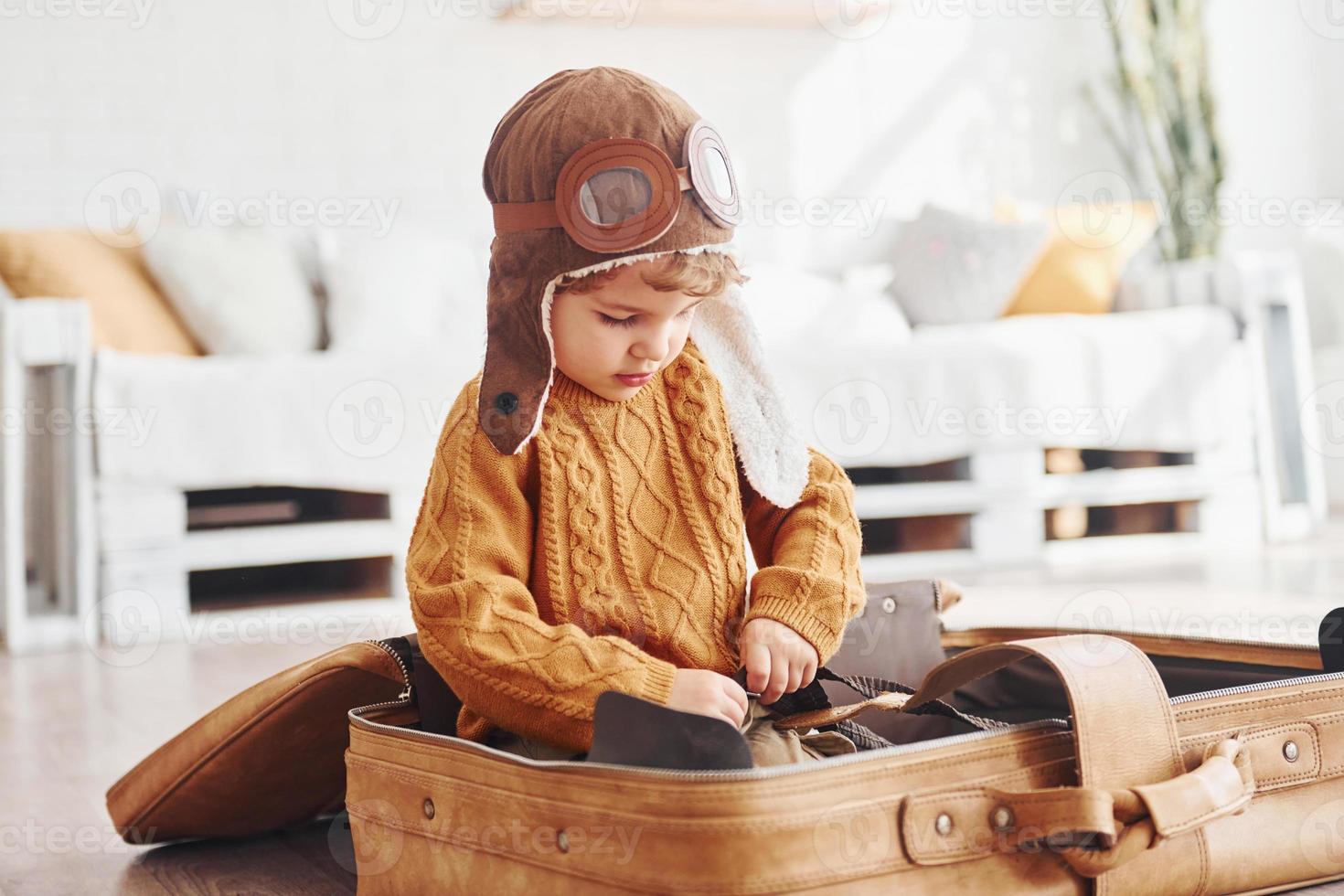 los niños pequeños con traje de piloto retro se divierten y se sientan en una maleta en el interior durante el día foto