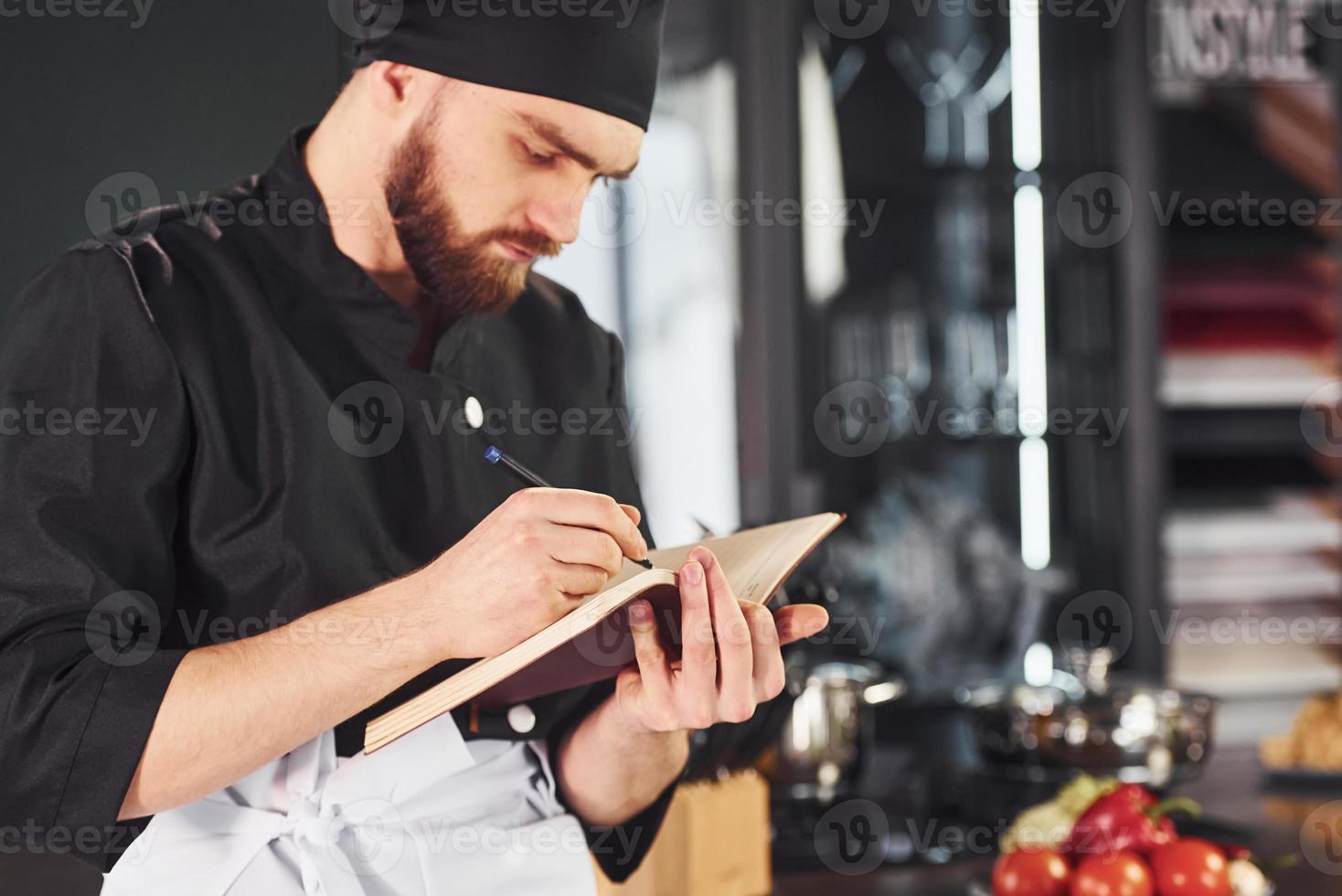 Cocinero profesional joven en uniforme de pie con bloc de notas en la cocina foto