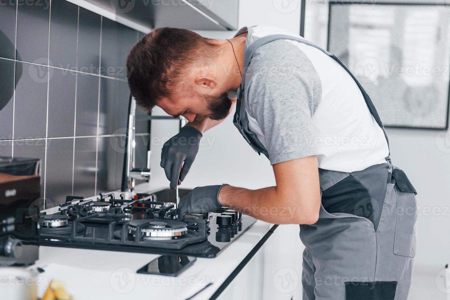 Young professional plumber in grey uniform working on the kitchen photo