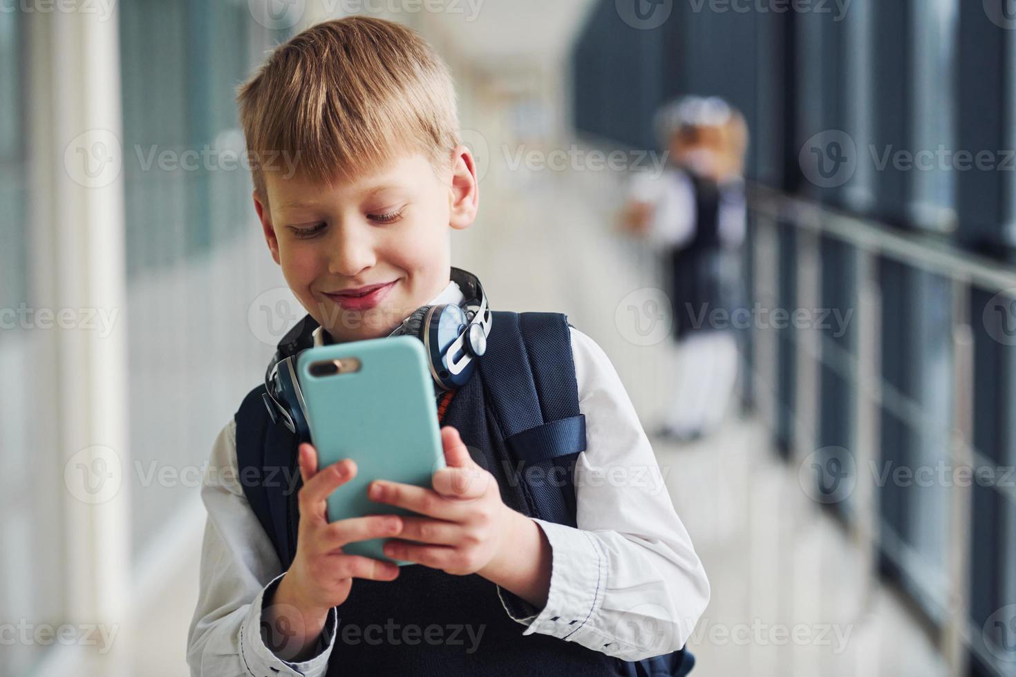 Little boywith phone and headphones standing if front of school kids in uniform that together in corridor. Conception of education photo