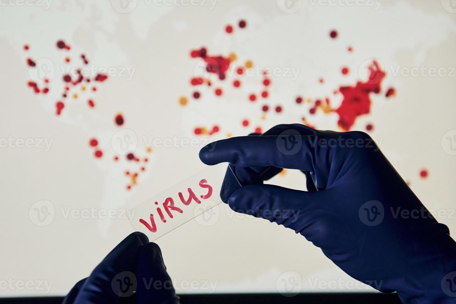 Man's hand holds lens with virus title on it against background with world map of infection photo