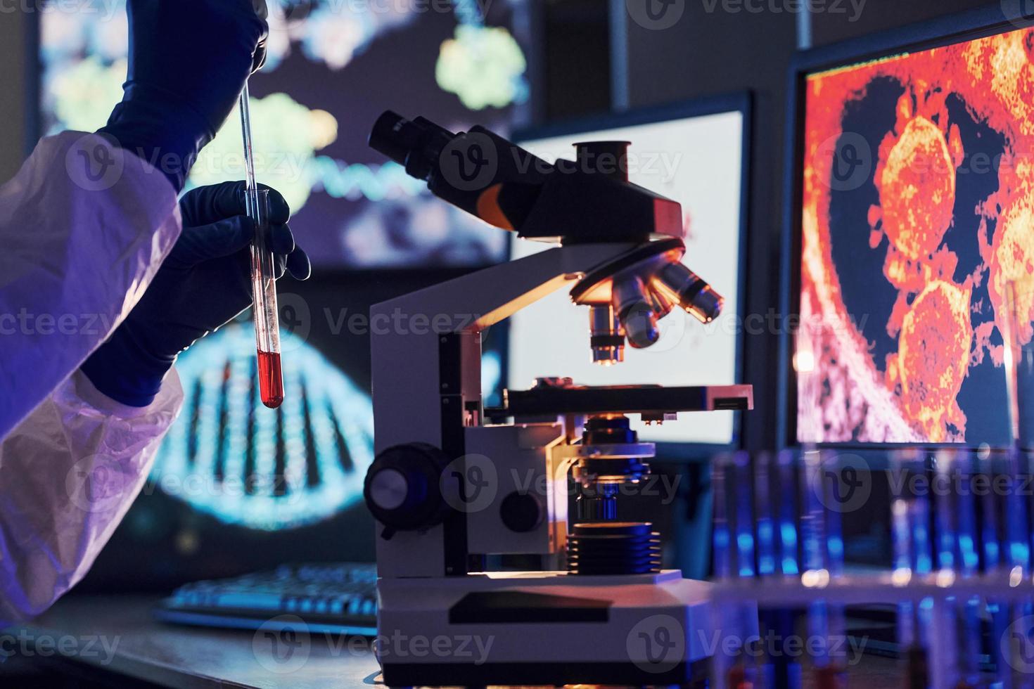 monitores con información sobre la mesa. científico con uniforme protector blanco trabaja con coronavirus y tubos de sangre en laboratorio foto