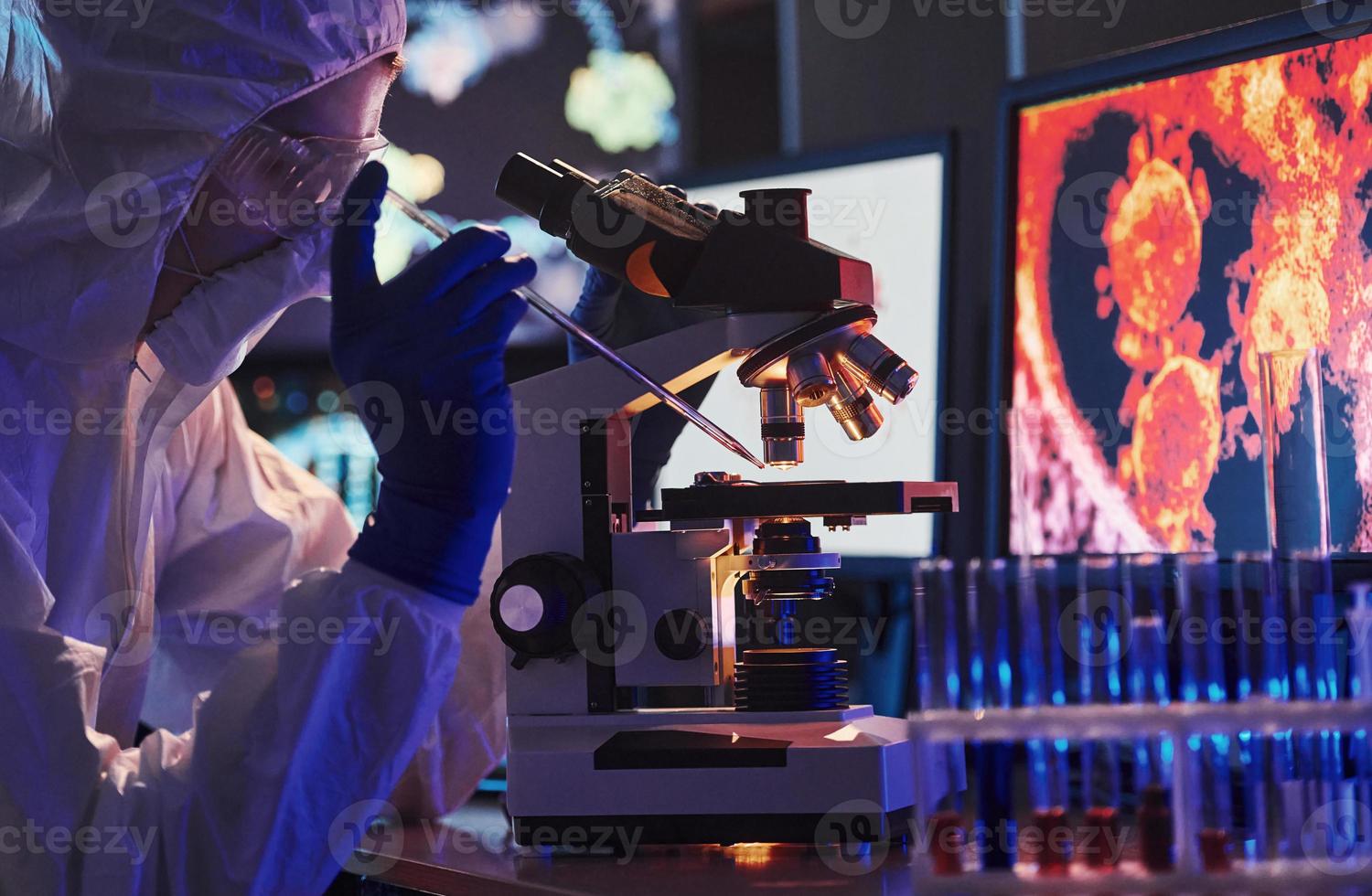 científico con uniforme protector blanco cerca de pantallas con datos trabaja con coronavirus y tubos de sangre en laboratorio foto