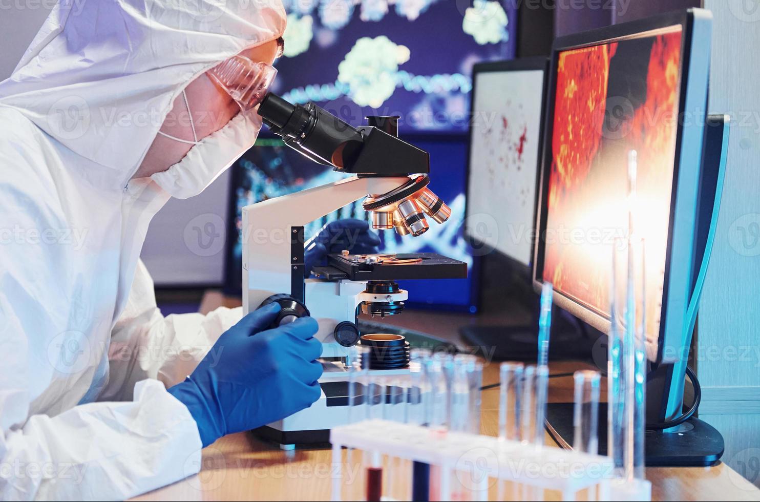 científico con uniforme protector blanco cerca de pantallas con datos trabaja con coronavirus y tubos de sangre en laboratorio foto